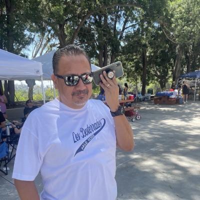 Picture of Eddie Santillan listening to something on his phone. He wears sunglasses and a Los Desterrados tshirt.